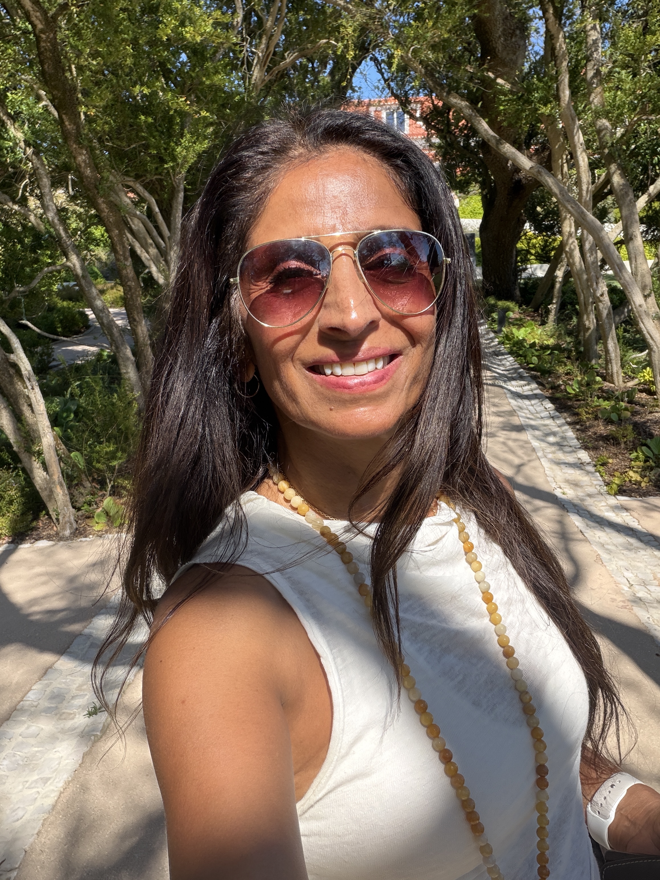 Smiling woman with long dark hair wearing aviator sunglasses, a white sleeveless top, and a beaded necklace outdoors on a sunny day with trees and a pathway in the background.