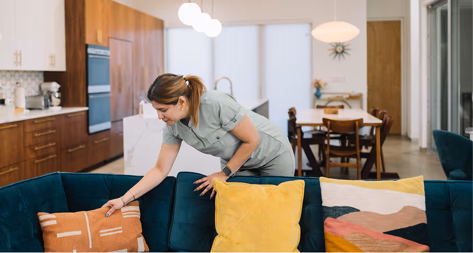 Woman tidying cushions on a dark teal sofa in a modern open-plan living and dining room.