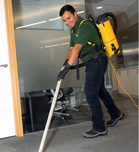 Man wearing a green shirt using a yellow backpack vacuum cleaner on a gray carpet in an office.