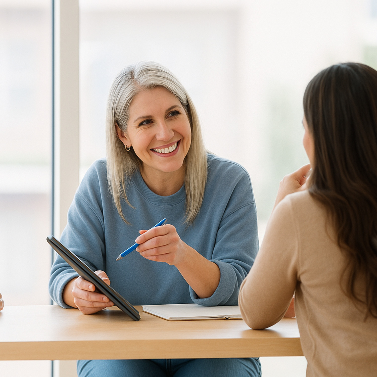 Smiling woman in a blue sweater holding a pen and tablet, speaking with another woman across the table in a bright, modern office setting.