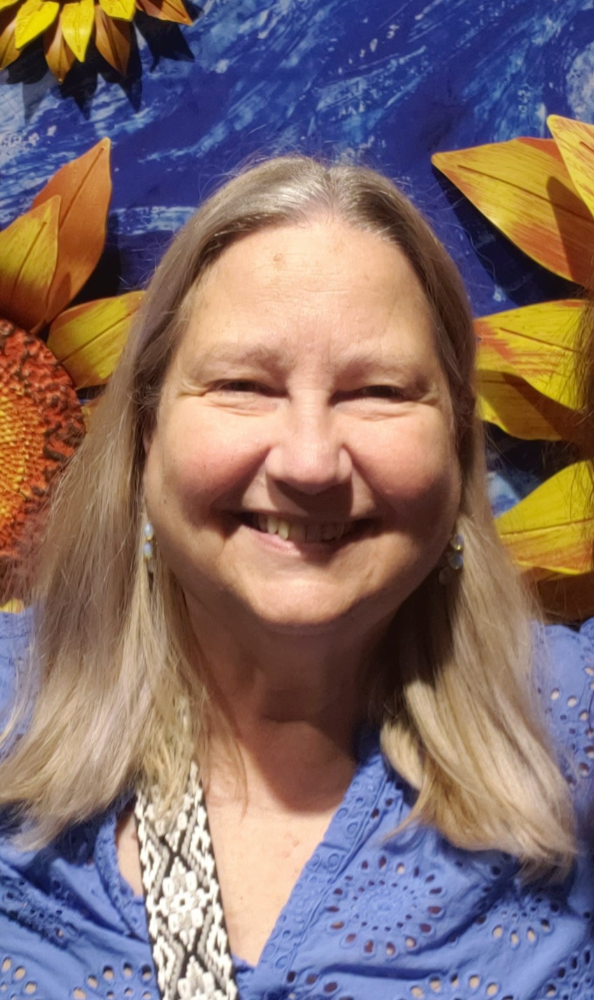 Laura Kemp: Smiling woman with long, light hair wearing a blue blouse, standing in front of a colorful sunflower-themed background.