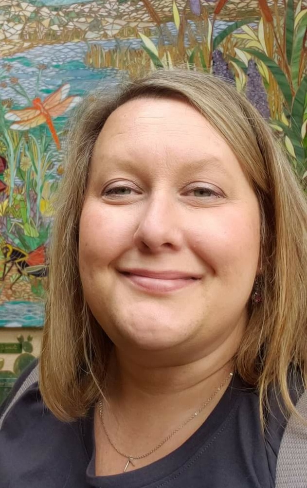 Nina Ballinger: Smiling woman with straight, blonde hair wearing a dark shirt, standing in front of a vibrant mosaic-style nature mural with flowers and dragonflies.