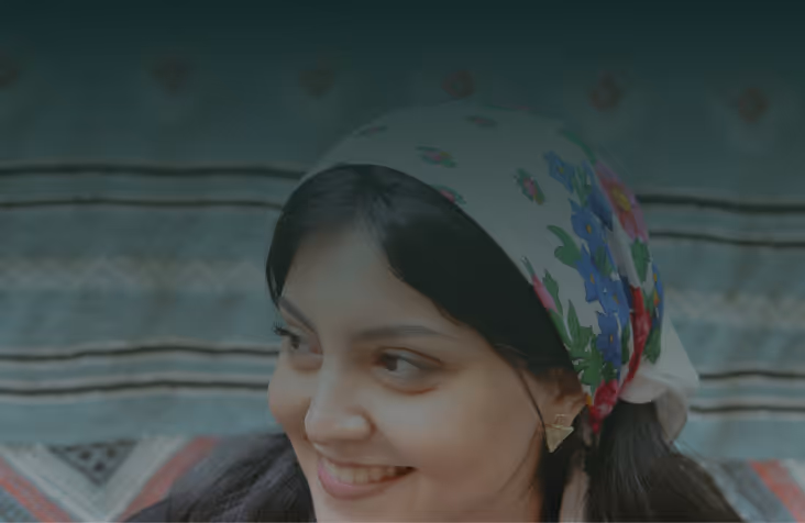 Smiling woman wearing a floral headscarf and gold earrings, looking to the side against a patterned background.