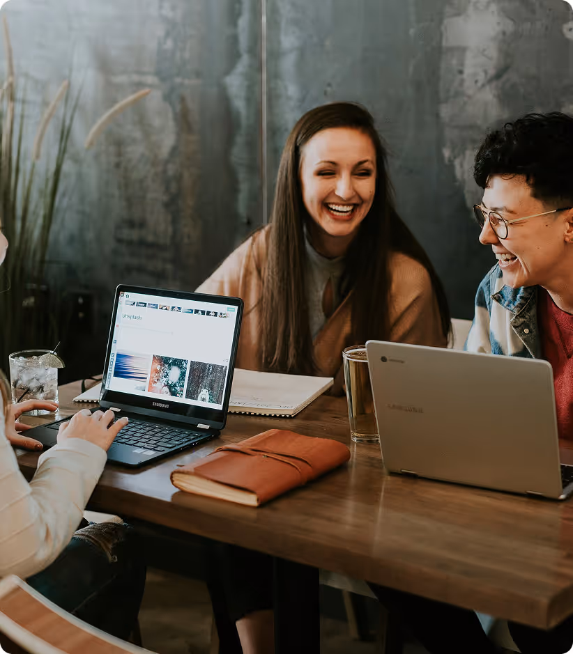 Three people sitting at a wooden table with laptops, laughing and engaging in a friendly conversation.