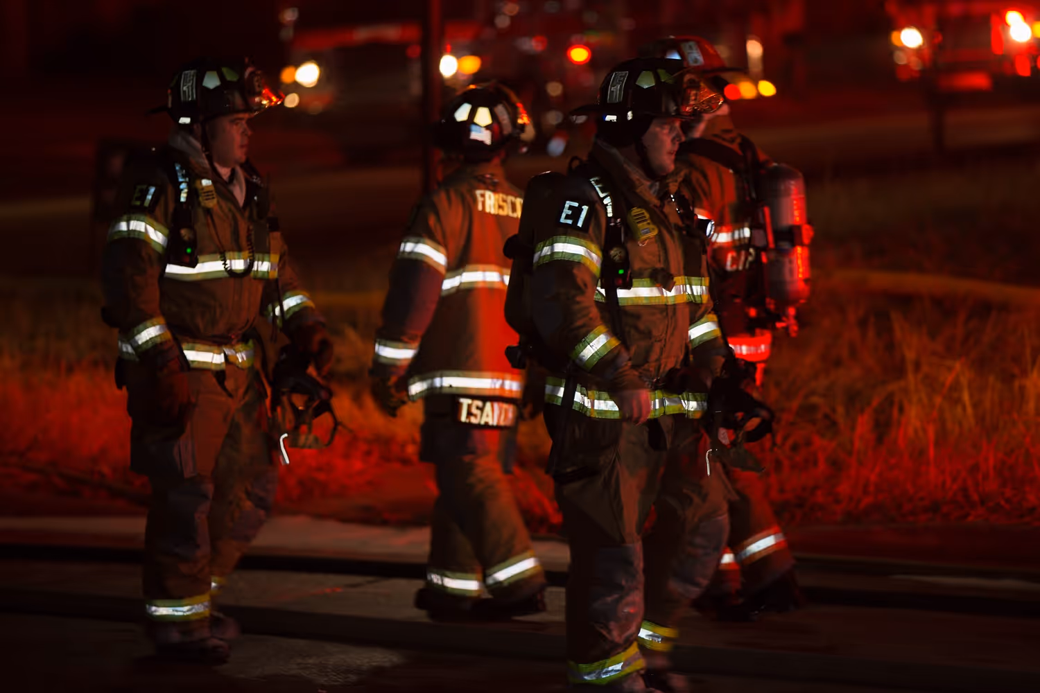 Group of firefighters in full turnout gear walking through a scene at night, lit by red emergency lights