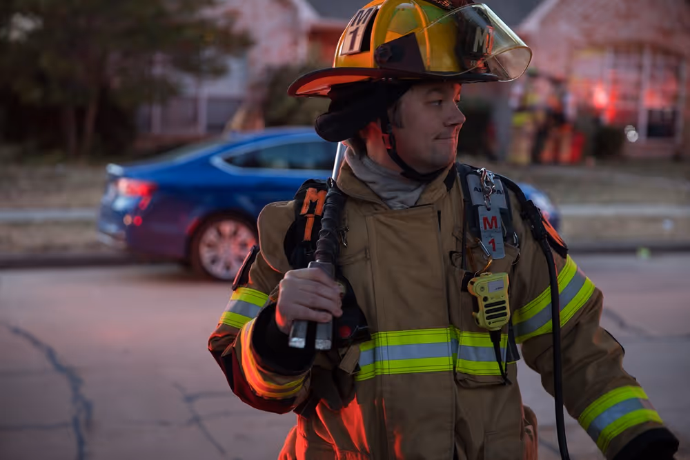 Frisco firefighter in full uniform holding equipment while standing on a residential street with emergency lights reflecting around him