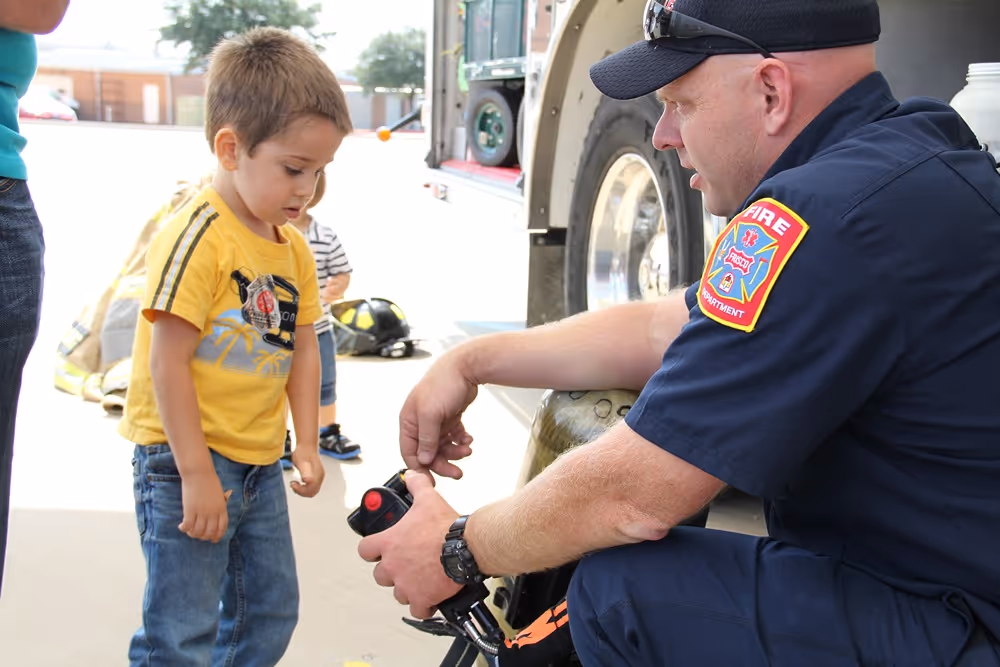 Frisco firefighter kneeling beside a young boy during a fire safety demonstration, showing him how equipment works