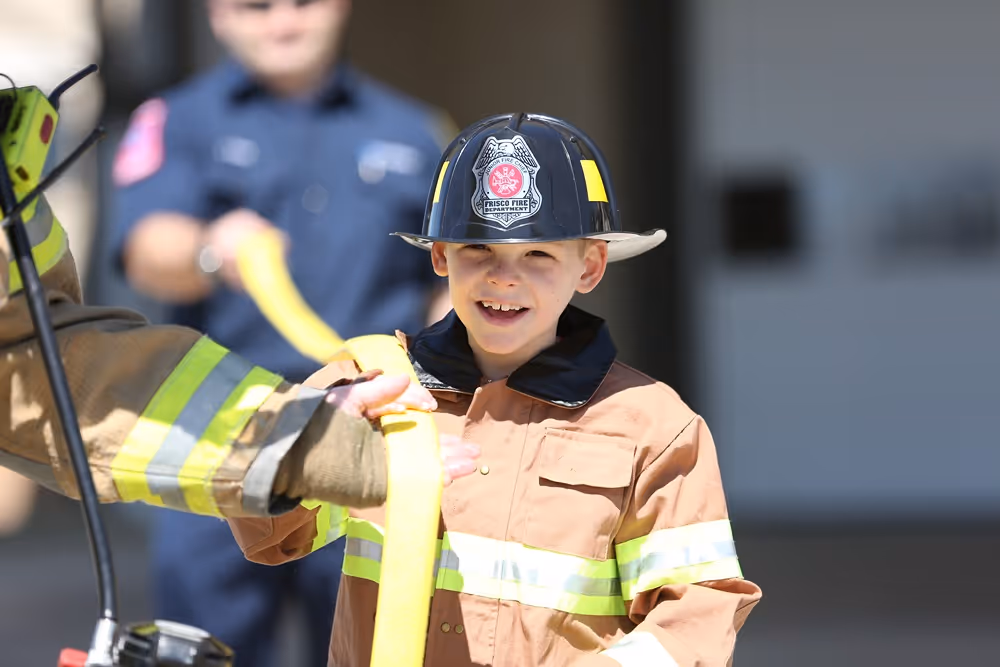 A young child dressed in firefighter gear holding a fire hose, with a firefighter in the background during a community event