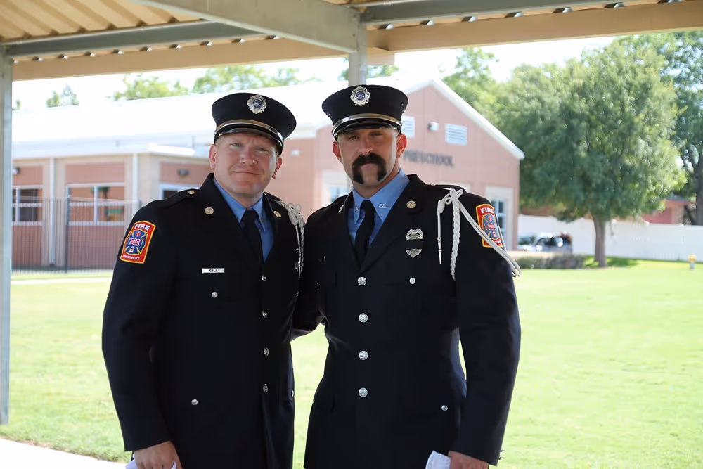 Two Frisco firefighters in formal dress uniforms posing together outside a fire station on a sunny day