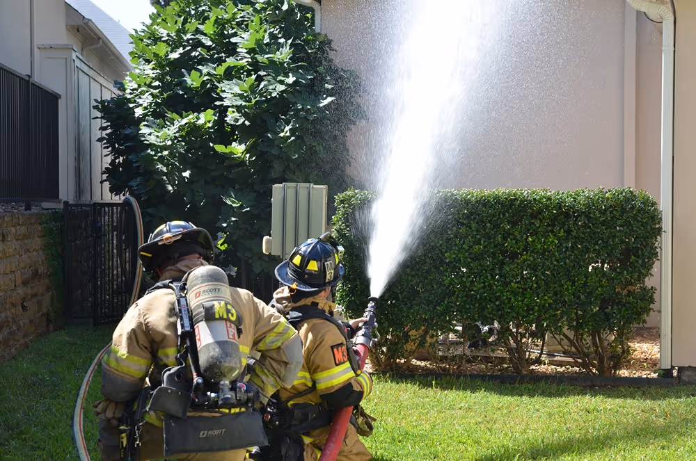 Two firefighters in full gear operating a hose and spraying water during a training exercise in a residential area
