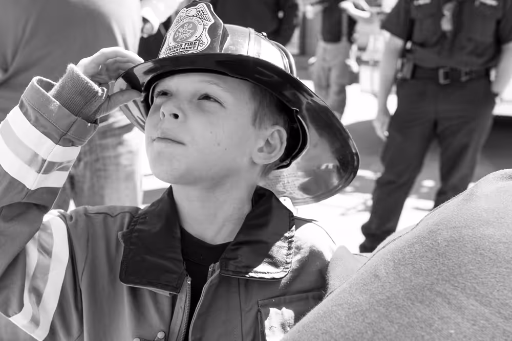 Young boy in firefighter uniform adjusting helmet during a community safety event, black and white photo