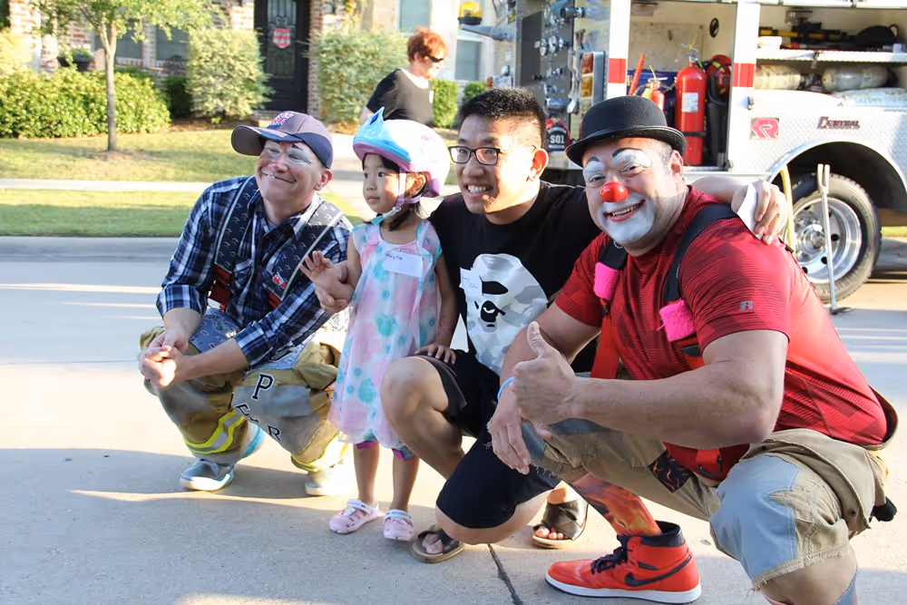 Firefighter, young girl, man, and a clown smiling for a photo during a community event on a sunny day