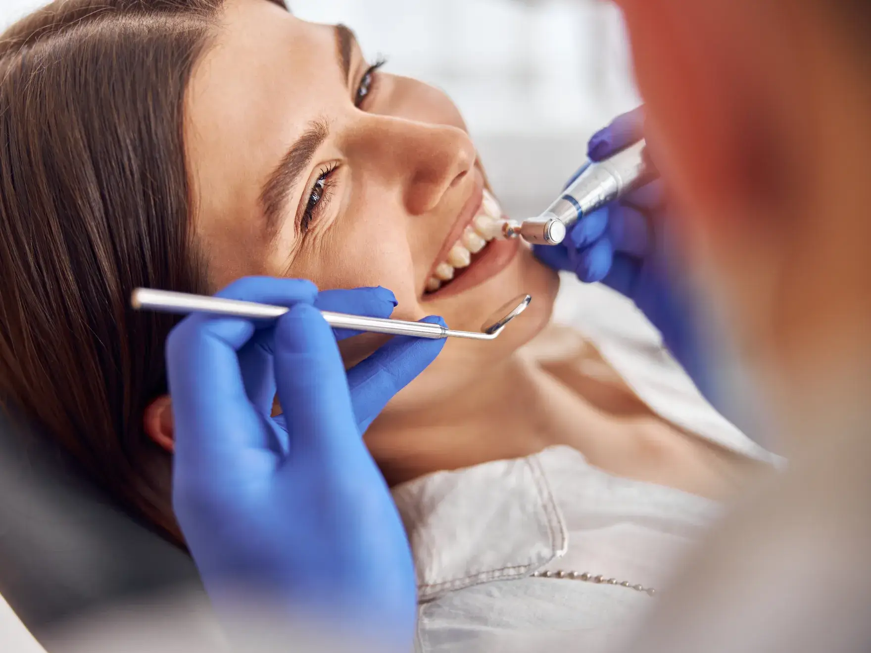 Dentist wearing blue gloves cleaning a woman's teeth with dental instruments.