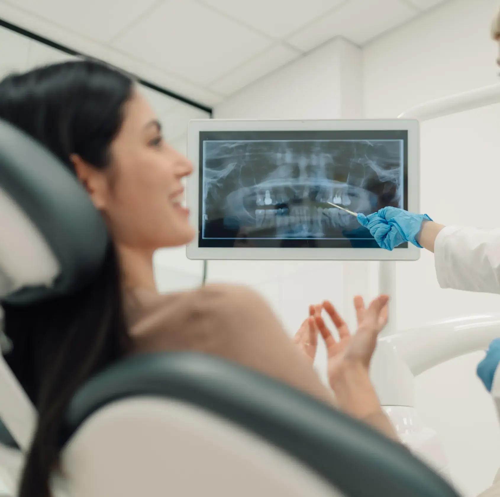Dentist with blue gloves pointing at a dental X-ray on a screen while a female patient sits in the dental chair smiling.