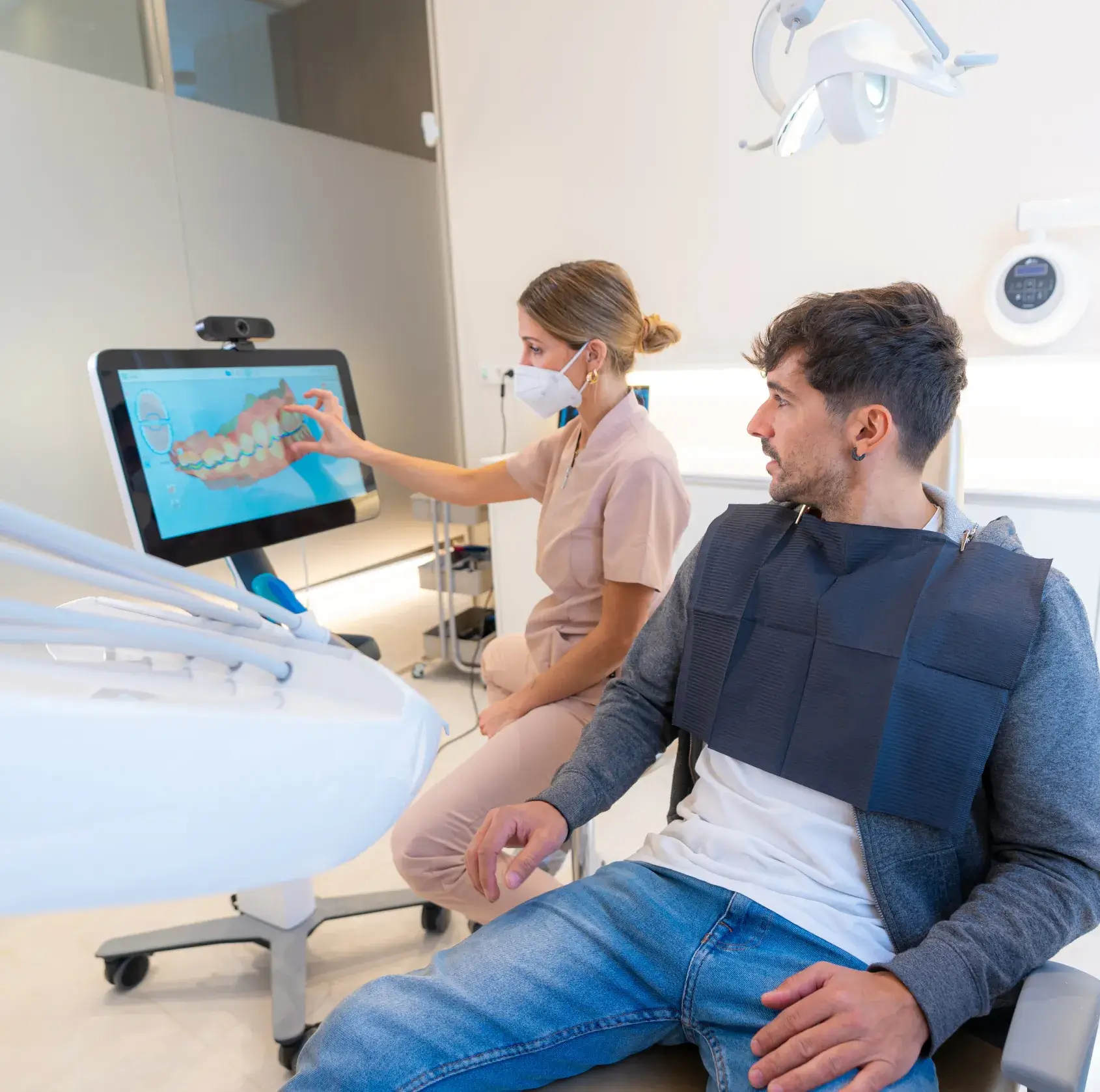 Dentist in scrubs and mask showing a digital 3D model of teeth on a screen to a male patient seated in a dental chair wearing a bib.