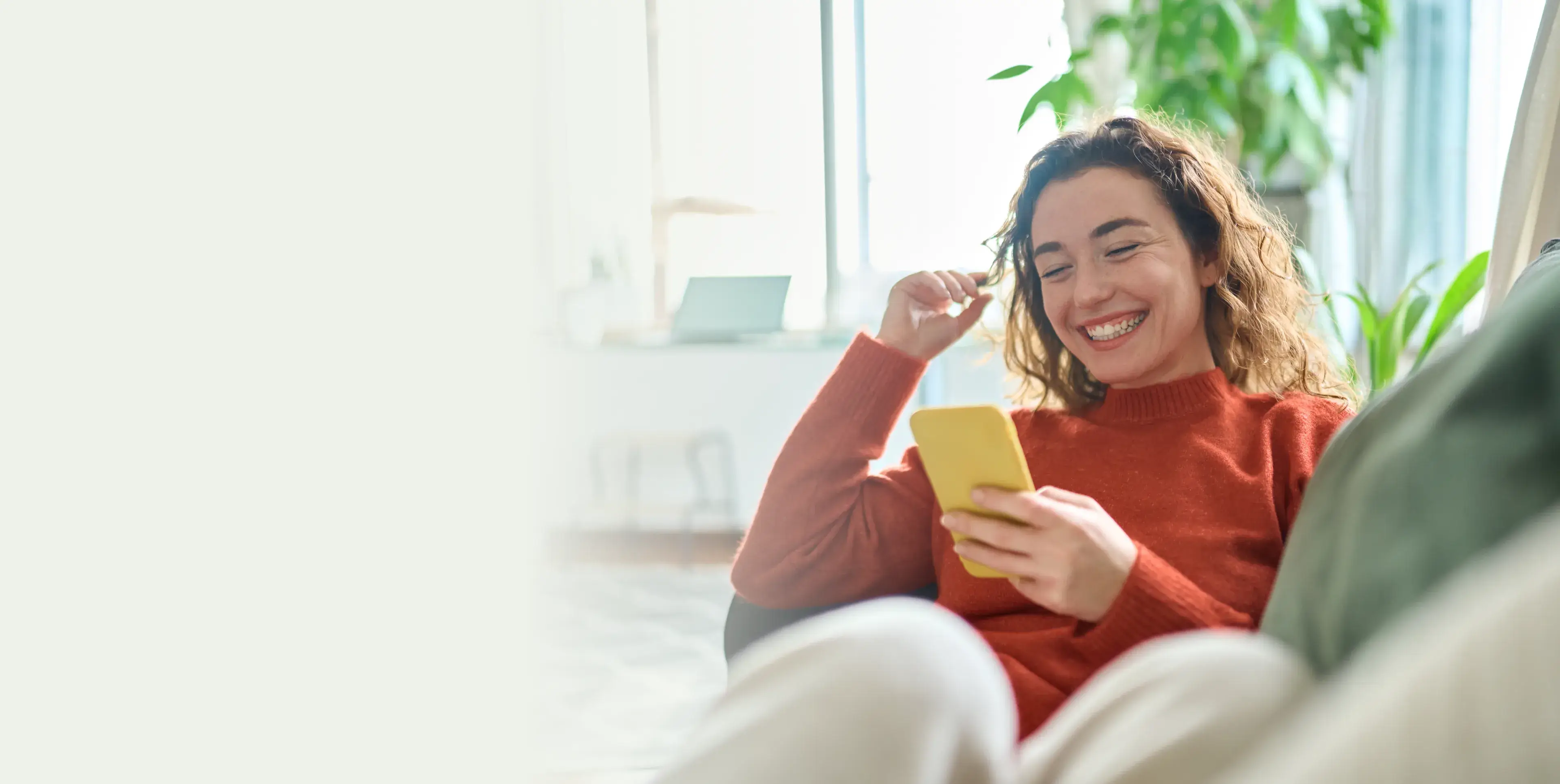 Smiling woman in a red sweater sitting comfortably and looking at a yellow smartphone in a bright room with green plants.