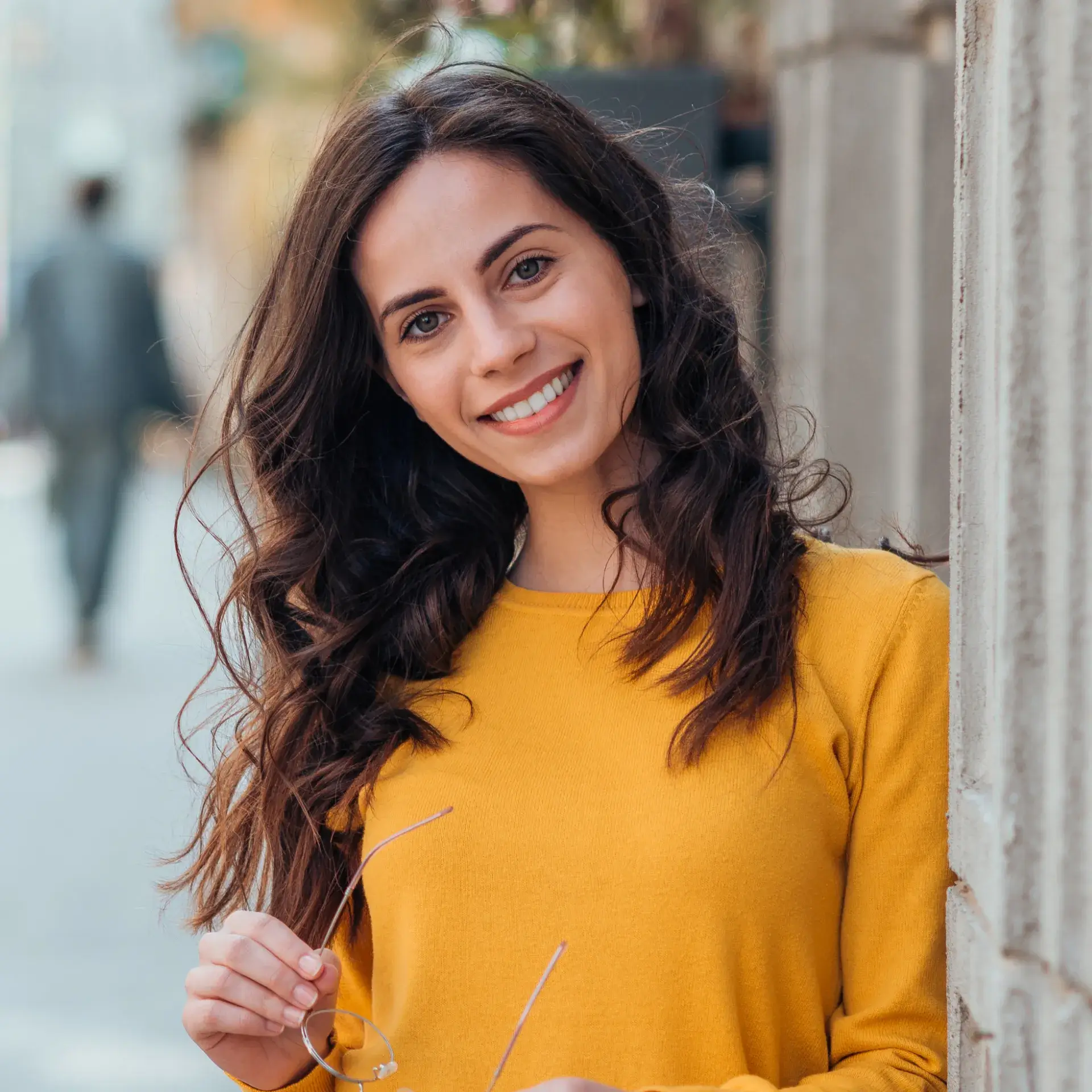Smiling woman with long curly hair wearing a yellow sweater and holding glasses, leaning against a wall outdoors.