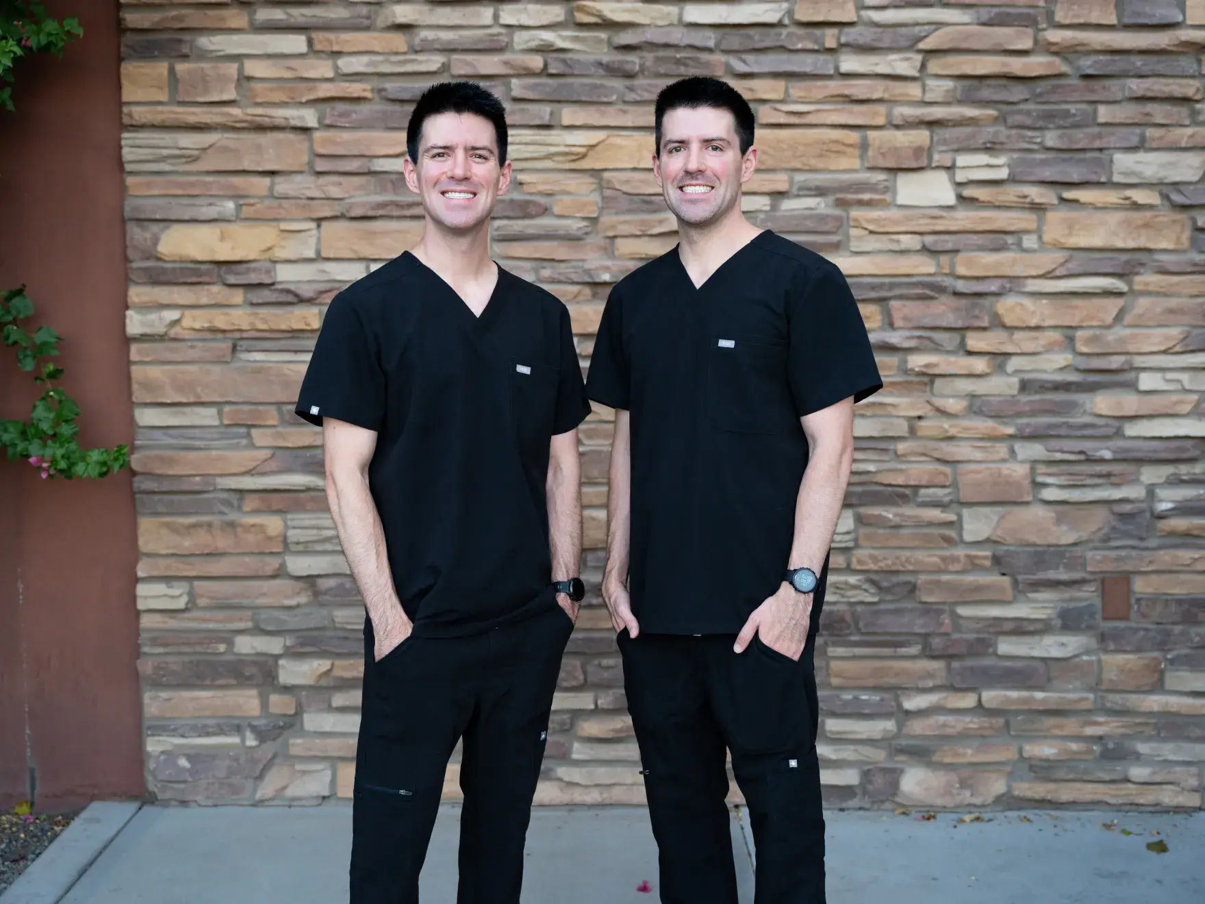 Two men wearing black medical scrubs standing and smiling in front of a stone wall.