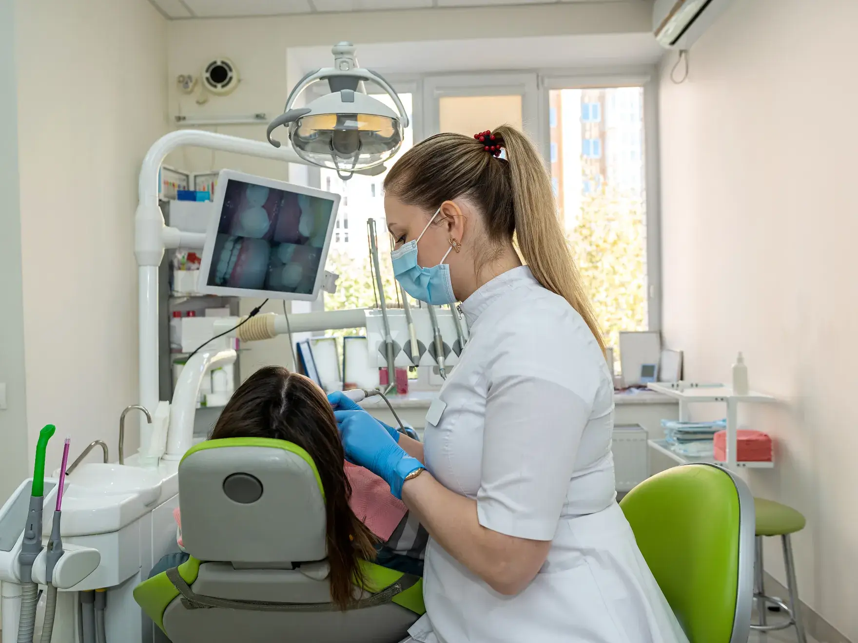 Dentist wearing a mask and gloves examining a patient's teeth in a dental office with dental equipment and a monitor displaying dental images.