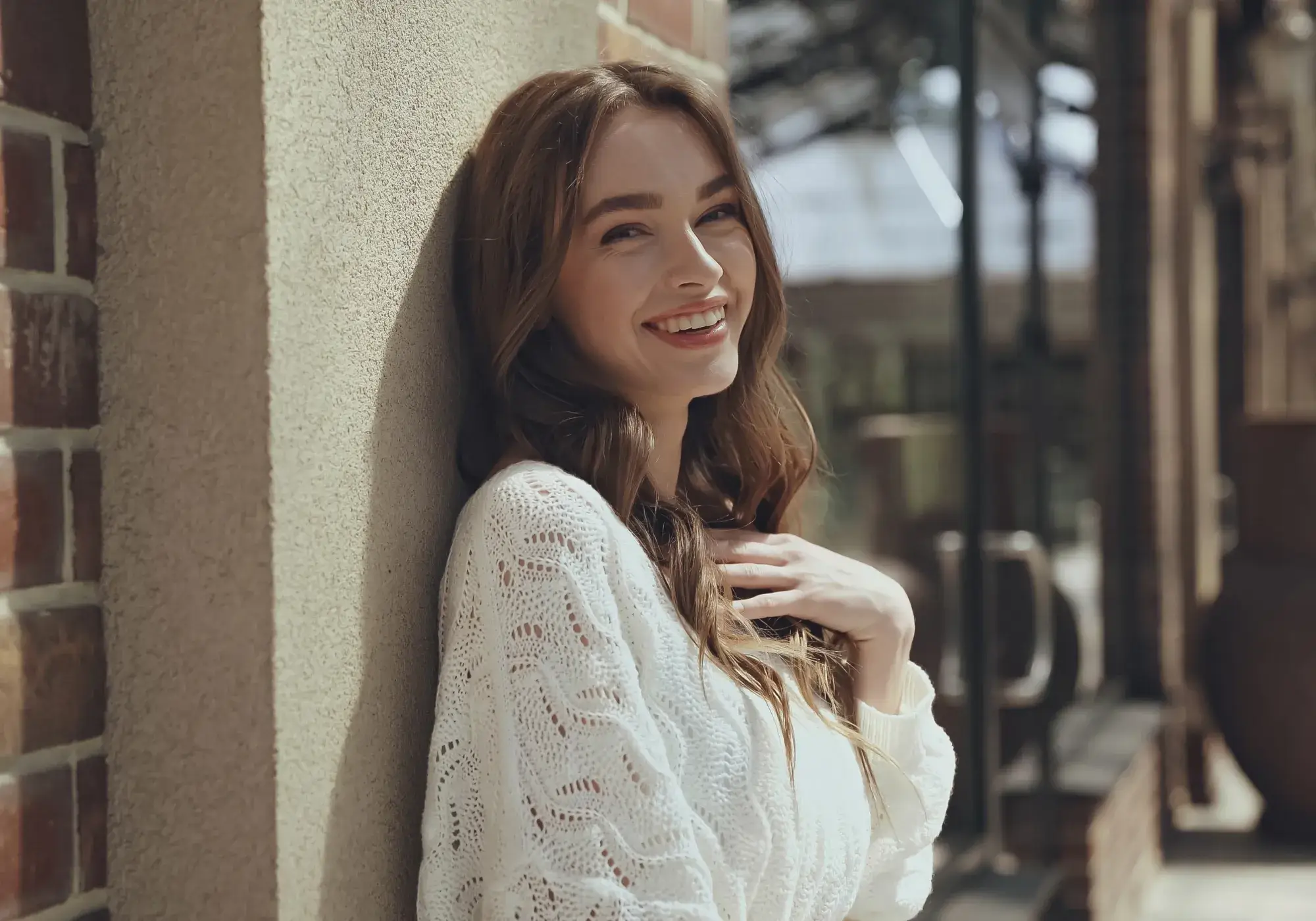 Young woman in a white knitted sweater smiling and leaning against a textured wall outdoors.