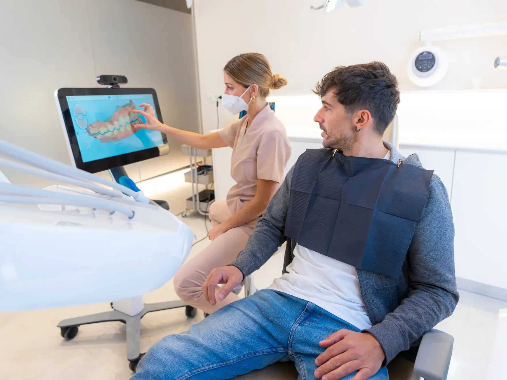 Dentist in scrubs and mask showing a dental scan on a screen to a seated male patient wearing a dental bib.