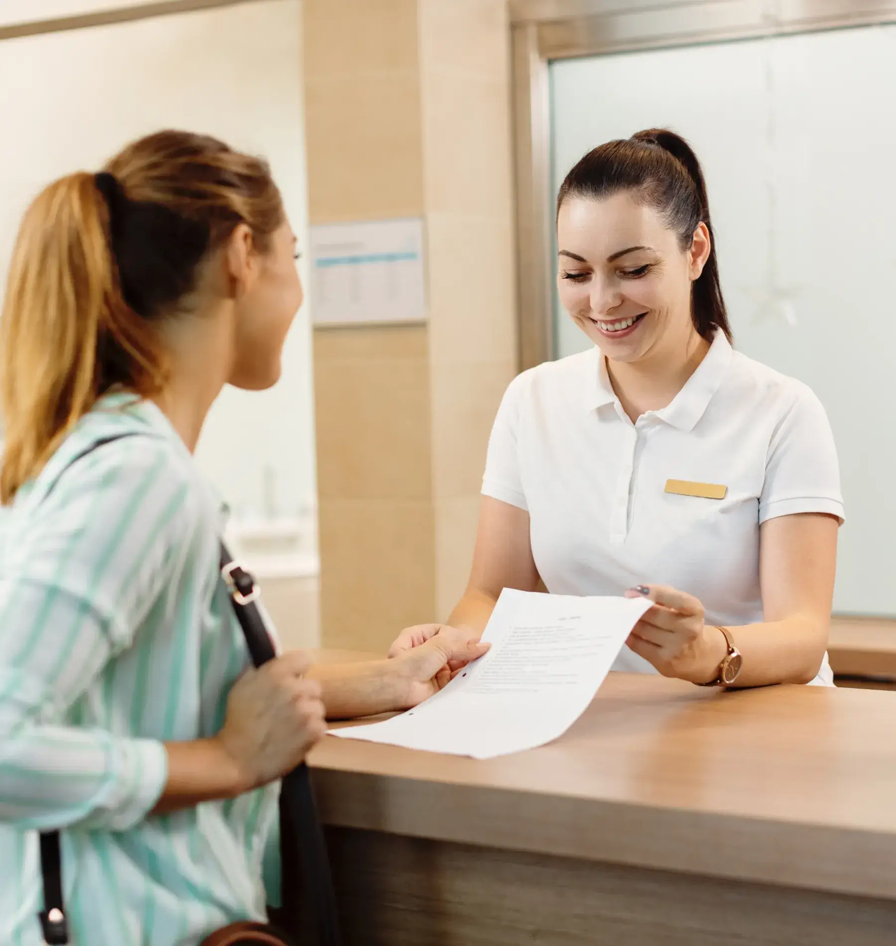 Smiling receptionist handing a document to a woman across a wooden desk in an office setting.