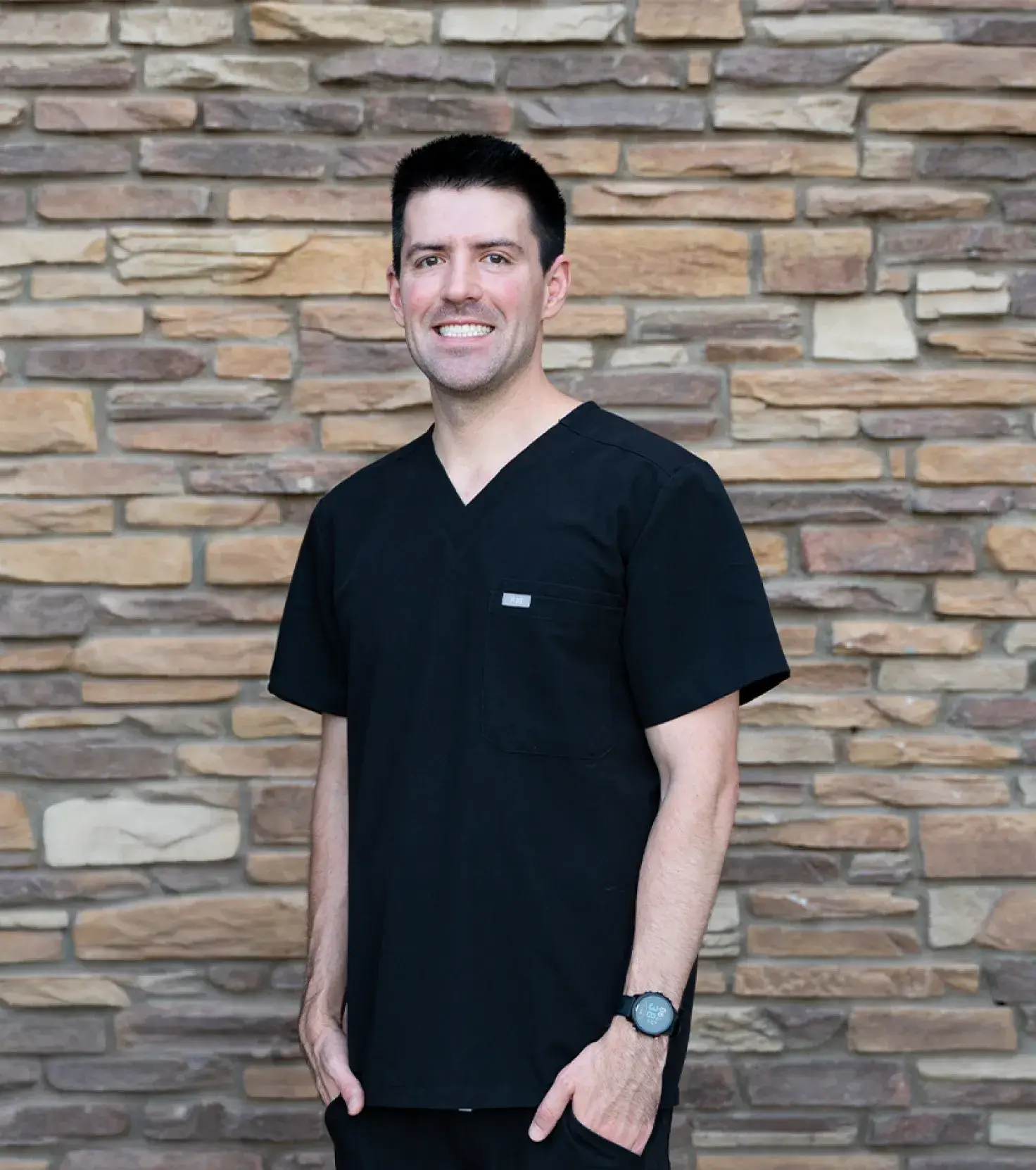 Smiling man wearing black medical scrubs standing in front of a stone wall.