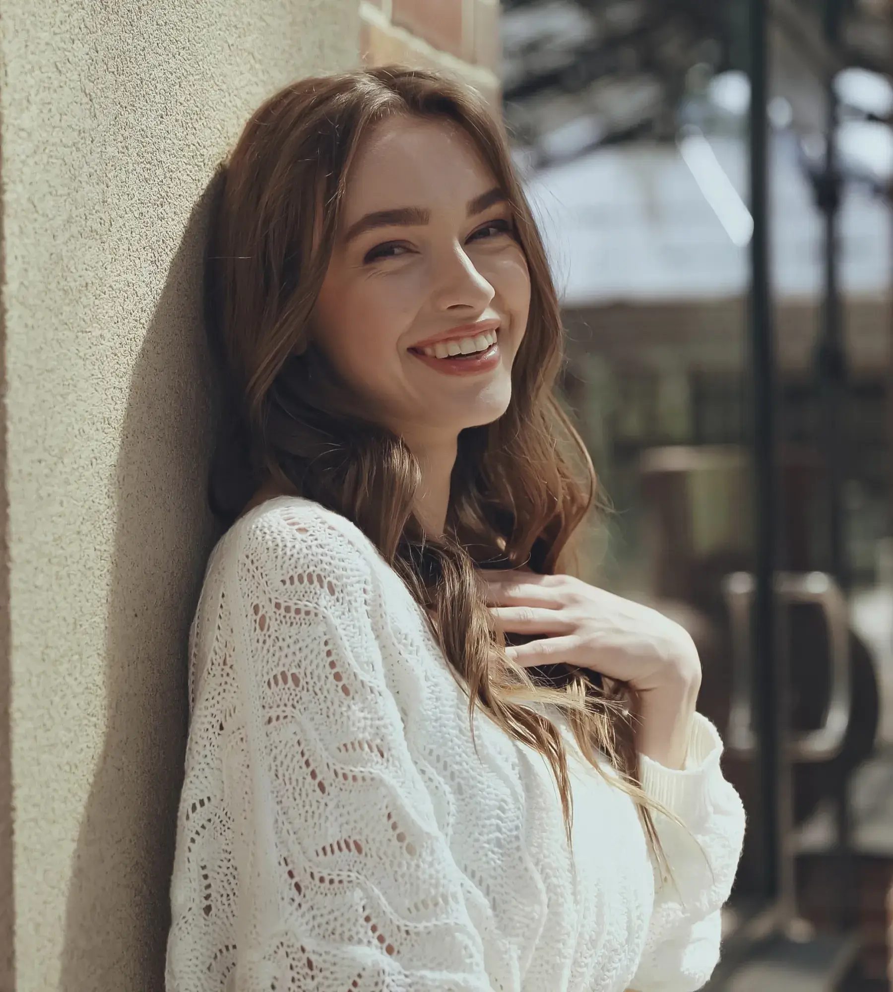 Smiling young woman with long brown hair leaning against a wall wearing a white knitted sweater.