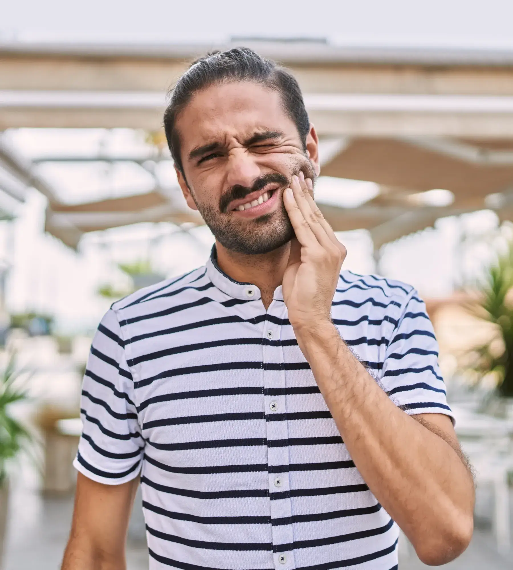 Man in a striped shirt holding his cheek with a pained expression, indicating toothache or jaw pain.