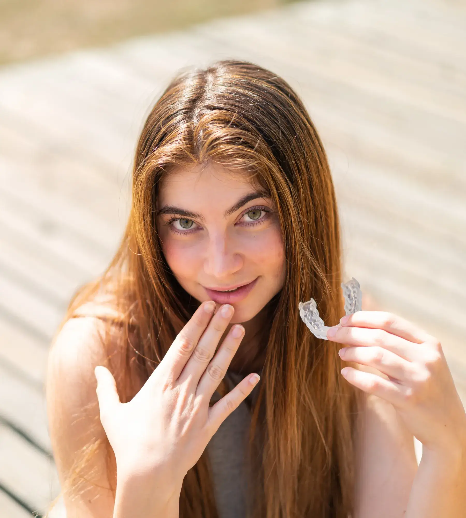 Young woman with long hair holding a clear dental aligner and covering her mouth with her other hand, smiling outdoors.