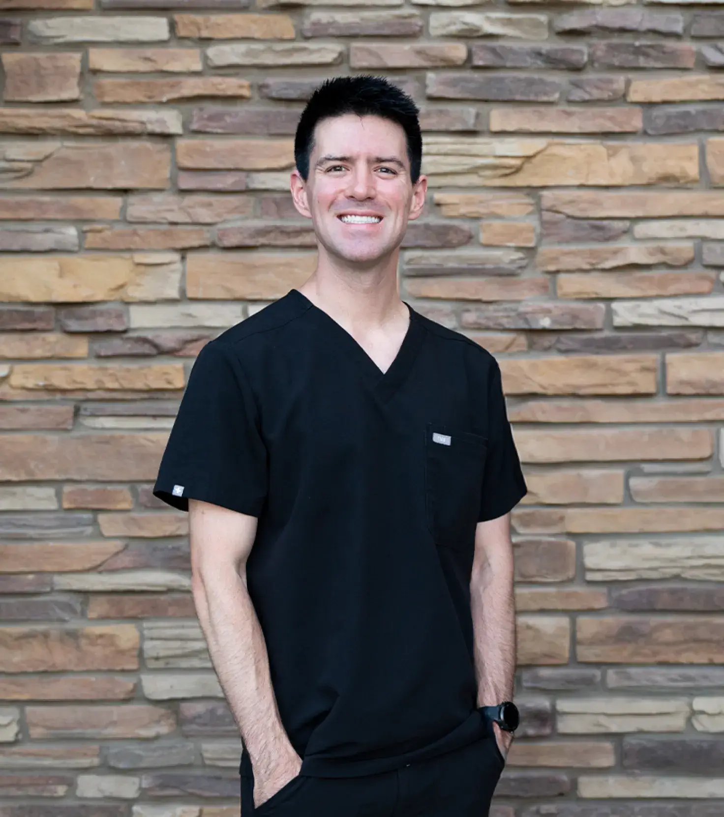 Smiling man wearing black medical scrubs standing in front of a stone brick wall.