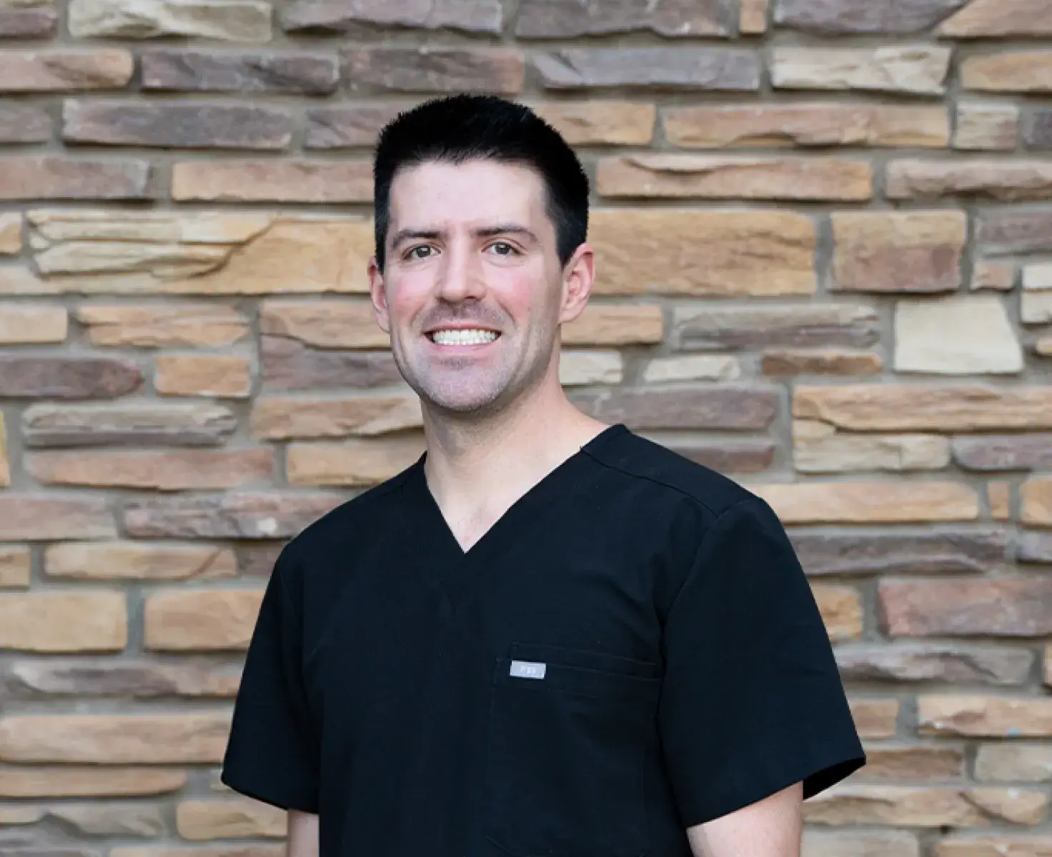 Smiling man with short dark hair wearing black scrubs standing in front of a stone brick wall.