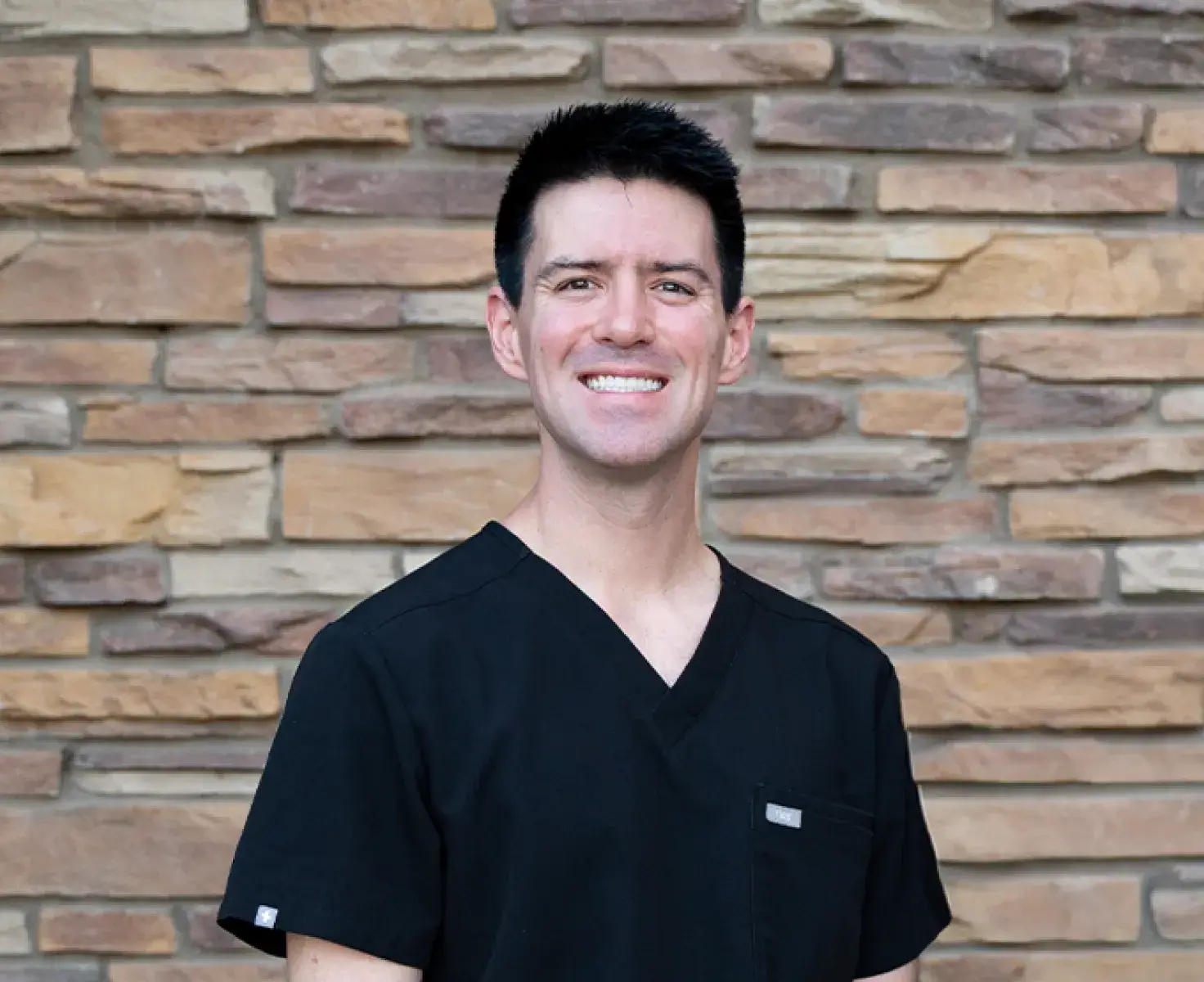 Smiling man wearing black medical scrubs standing in front of a stone brick wall.