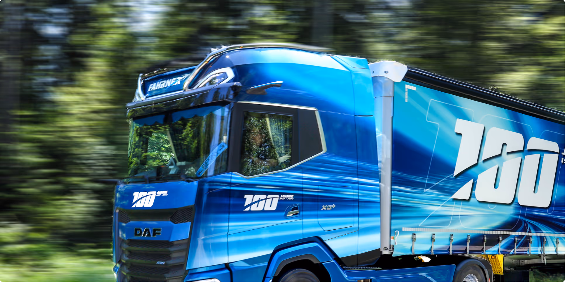 Moderner blauer LKW mit „100 Jahre“-Branding, fährt durch bewaldete Landschaft – Symbol für Innovation und Tradition im Transportwesen