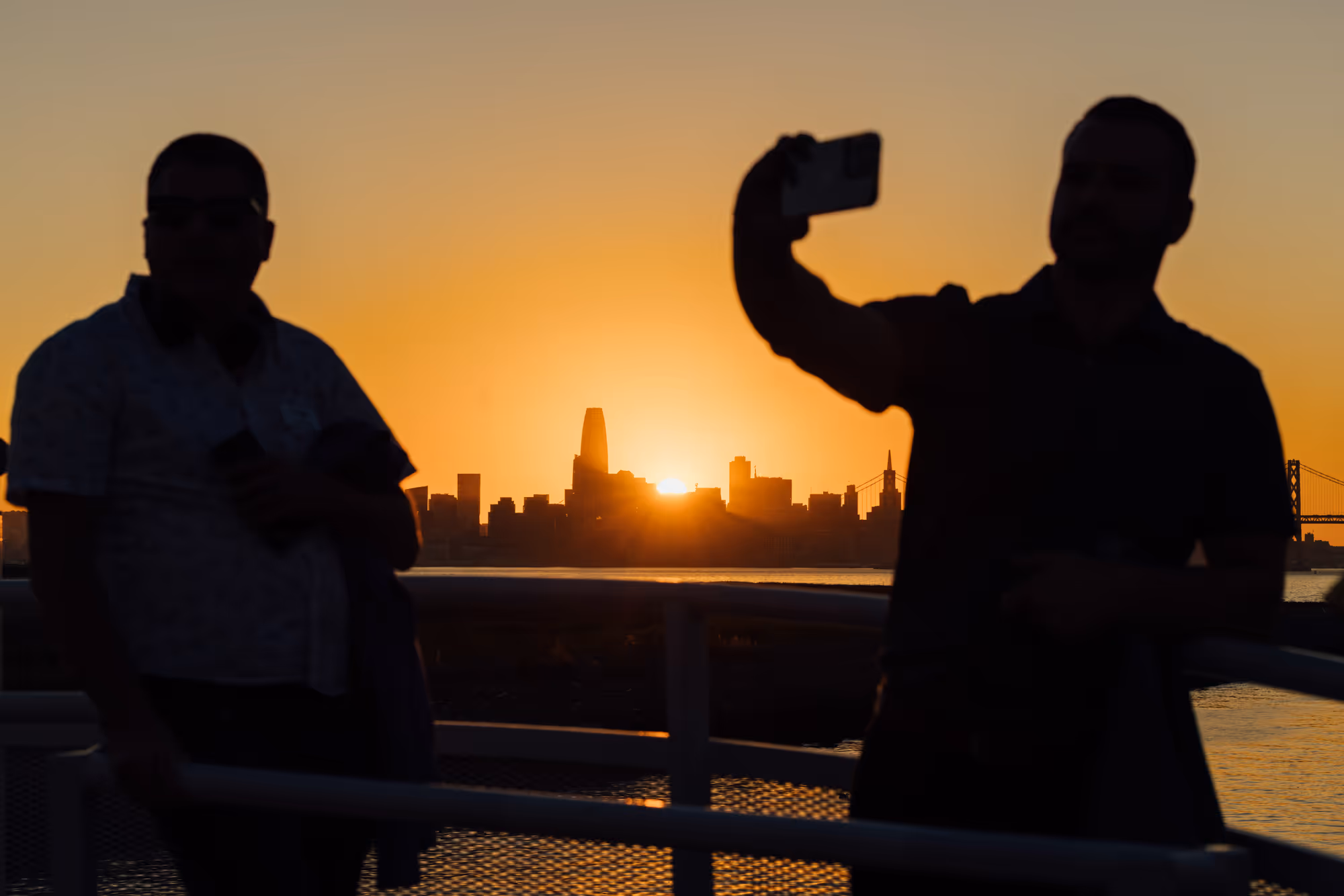 Silhouettes of two men by a railing with a smartphone, with the city skyline and setting sun in the background.