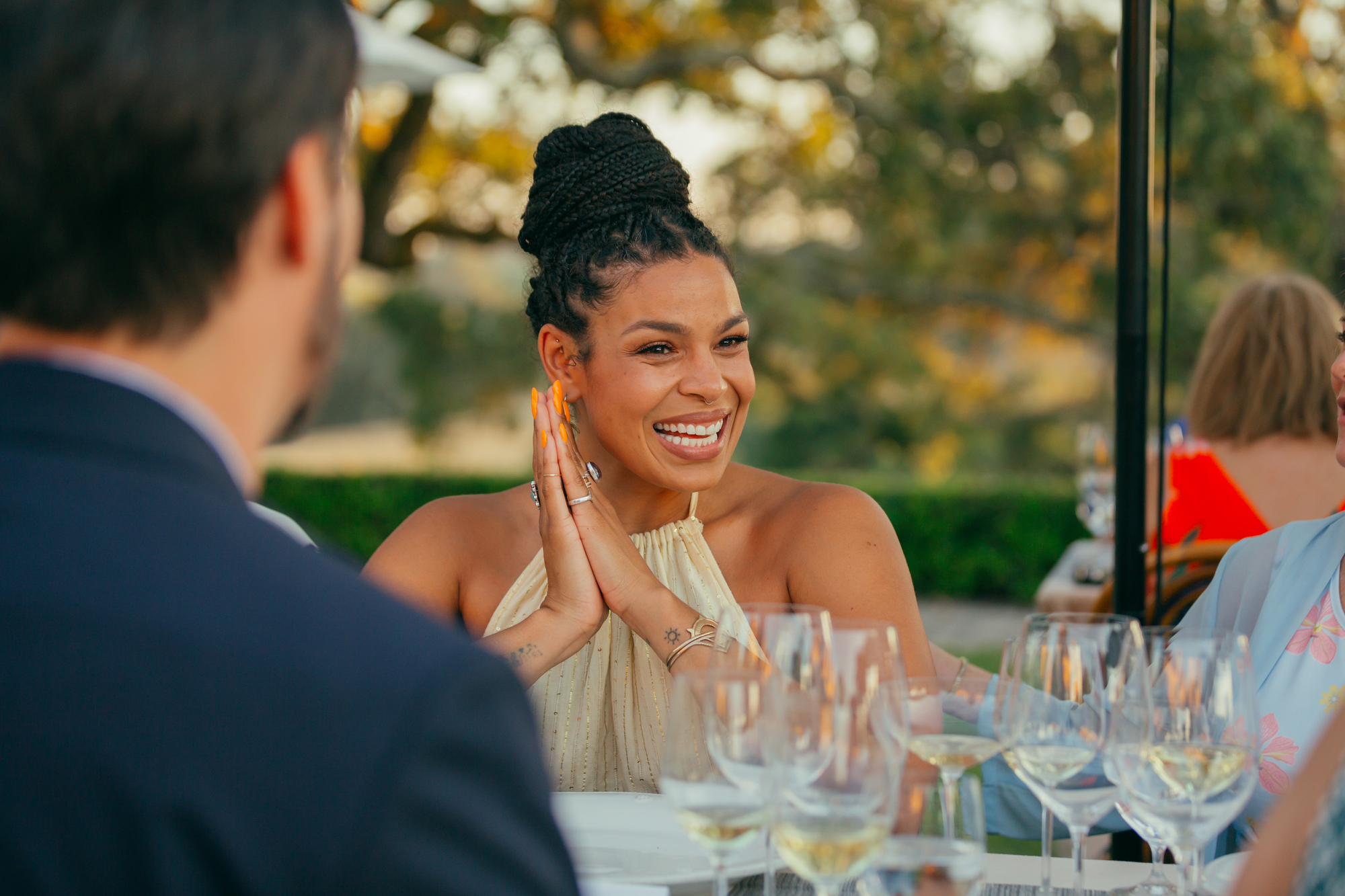 Smiling woman with braided hair and bright orange nails sitting at a table with wine glasses outdoors, engaging in conversation.