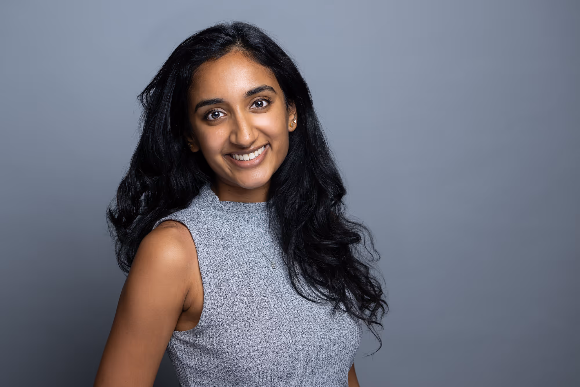 Smiling woman with long black hair wearing a sleeveless gray top against a gray background.