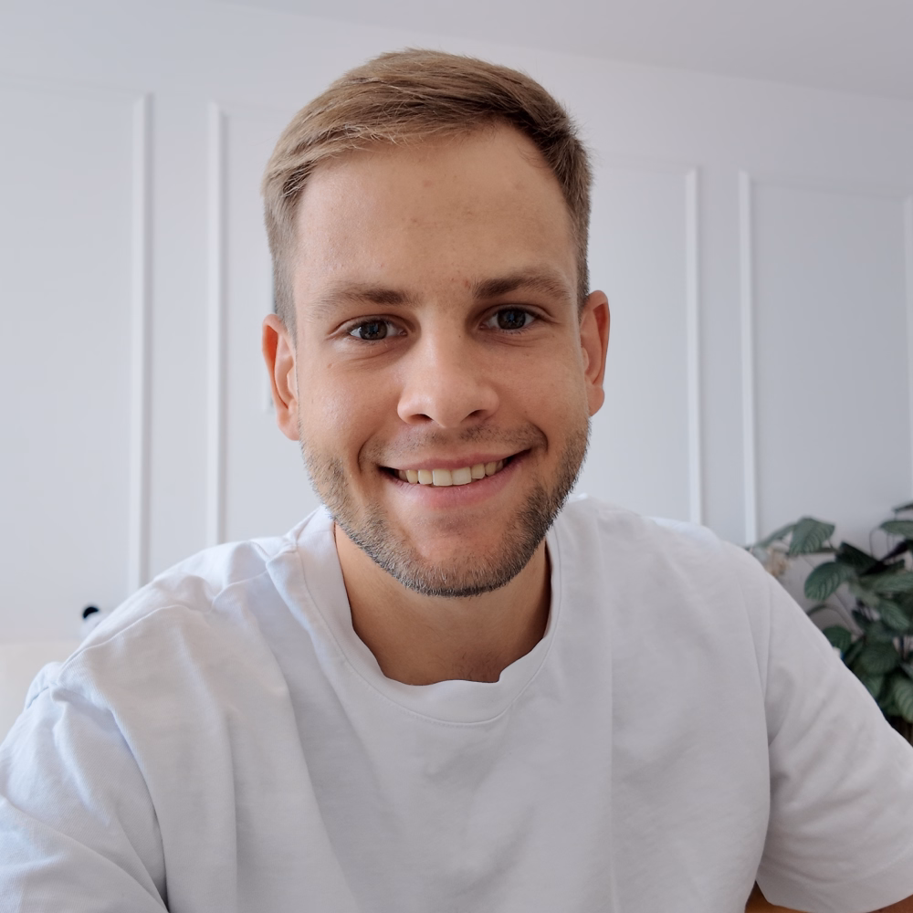 Smiling young man with short brown hair and beard wearing white t-shirt indoors.