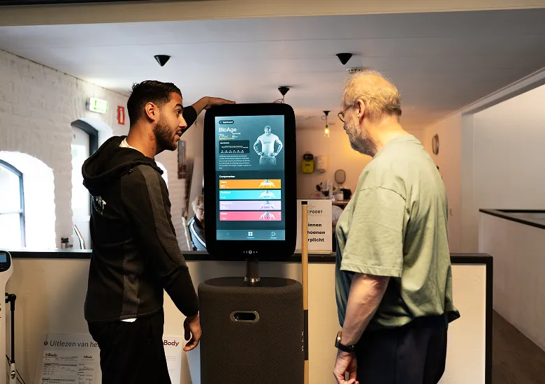 Een trainer van Fitpoort Healthclub geeft uitleg over de Fitness Hub aan een oudere man.