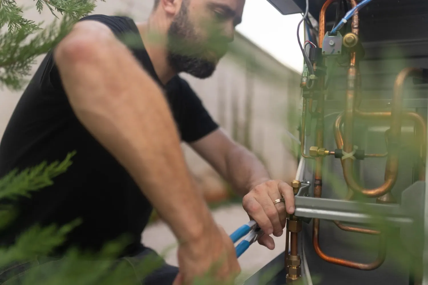 A man with a beard, seen through green foliage, uses pliers to work on copper pipes of an outdoor AC unit