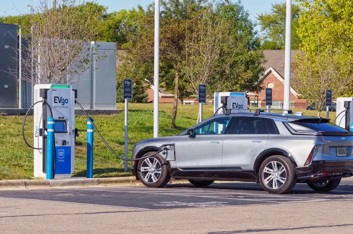 A silver Cadillac Lyriq is charging at an EVgo station with multiple charging units in an outdoor parking lot.