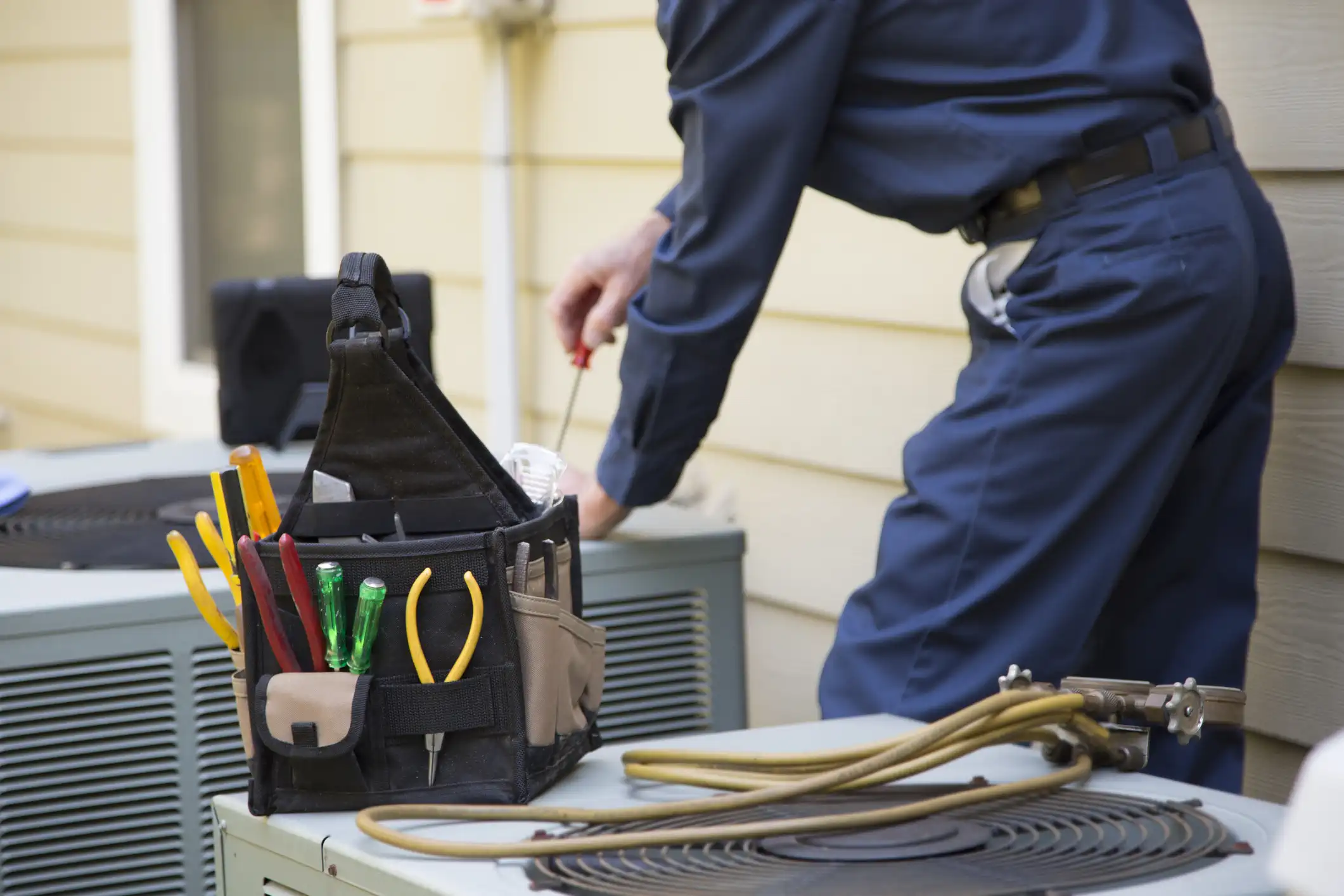 A close-up image focuses on a black and tan tool bag full of various tools, including screwdrivers and pliers, sitting on top of an outdoor air conditioning (AC) unit. In the background, an HVAC technician wearing a dark blue uniform bends over to service a second AC unit, holding a screwdriver. The coils, fan guard, and refrigerant lines of the AC unit in the foreground are visible.