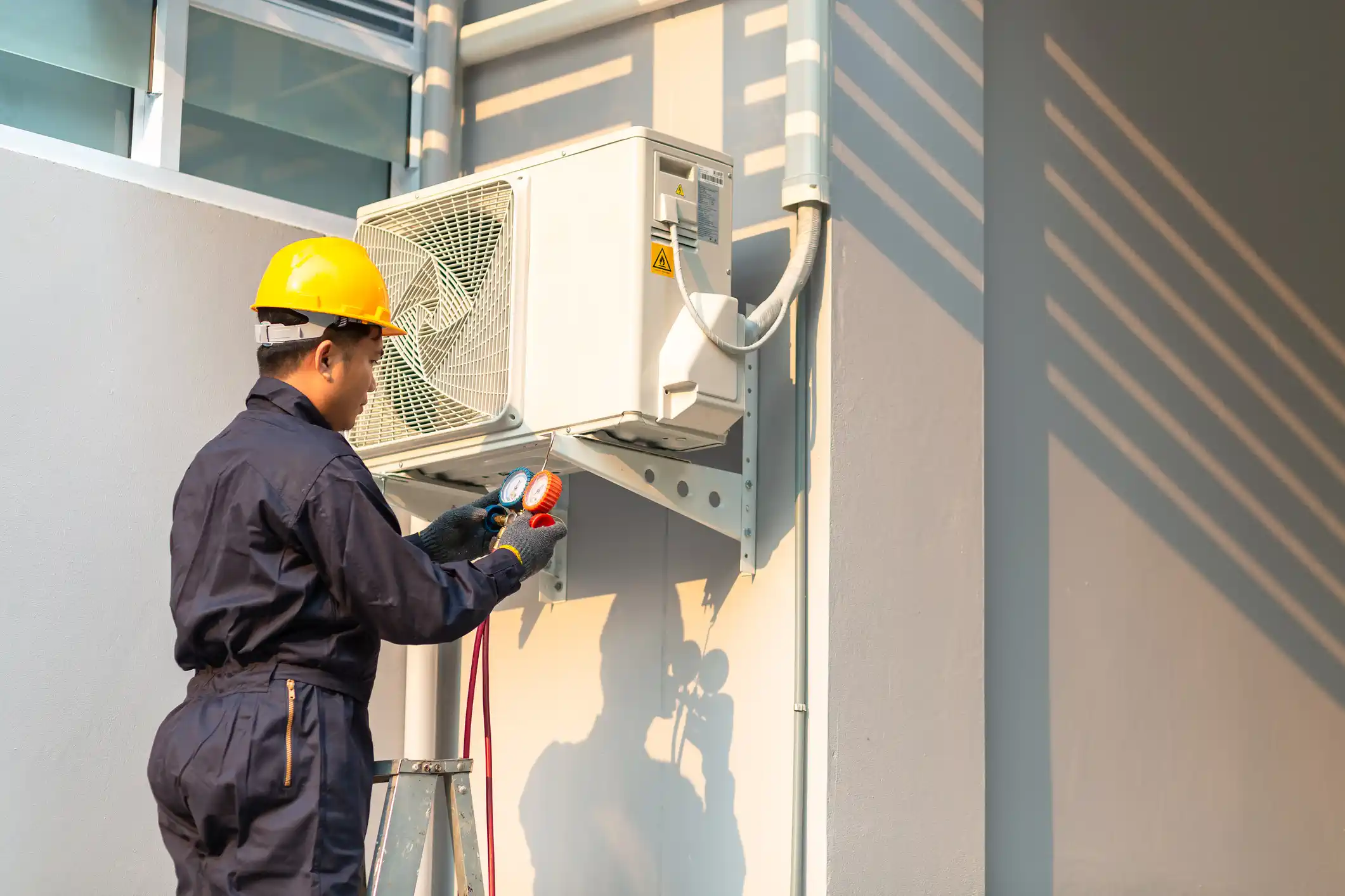 A technician, seen from the back, wearing a dark blue jumpsuit, a yellow safety hard hat, and protective gloves, is servicing a white wall-mounted outdoor AC unit