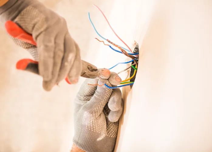 An electrician wearing gloves uses pliers to work on electrical wires coming out of a wall.