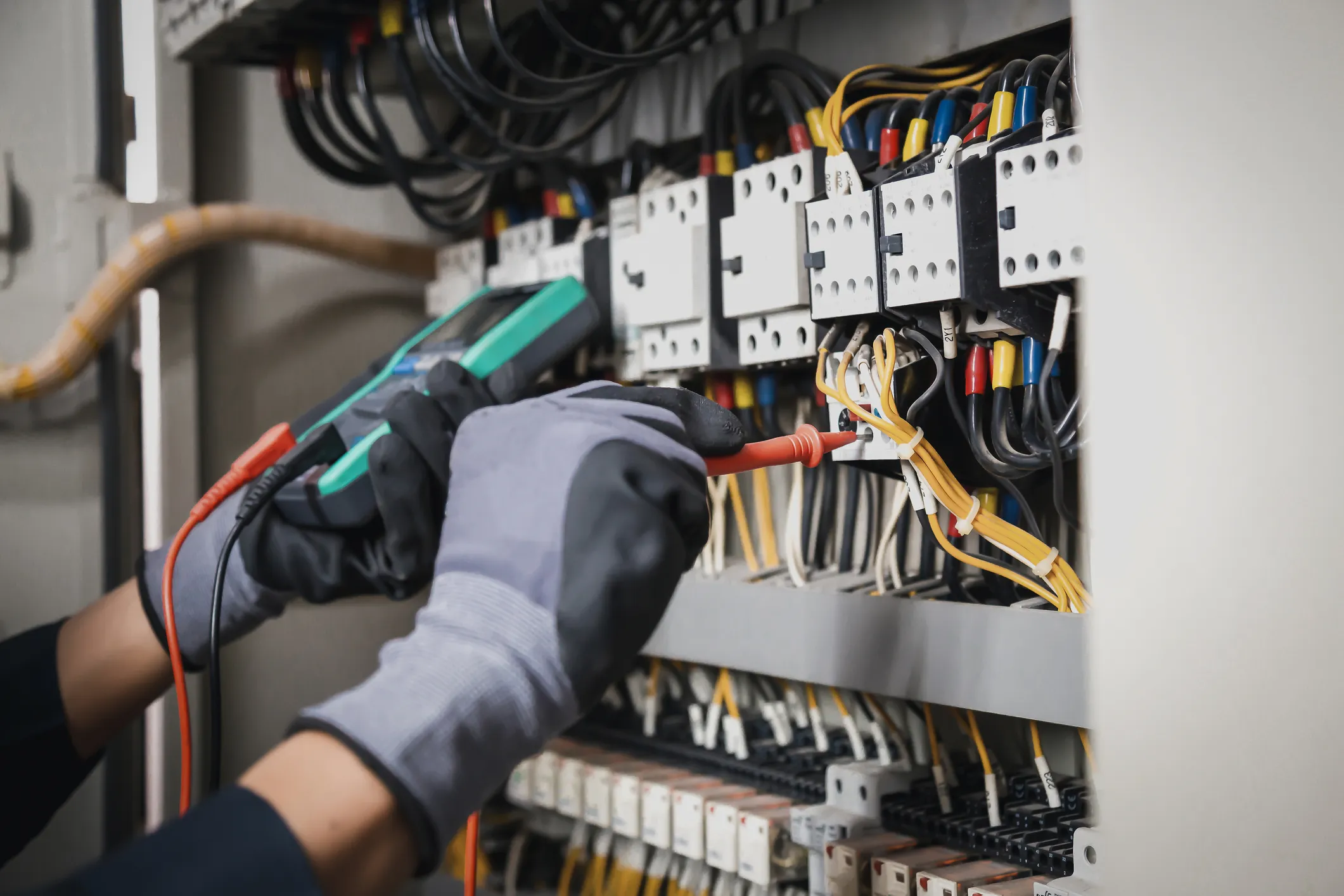 An electrician wearing gloves uses a multimeter to test the wiring inside an electrical control panel.