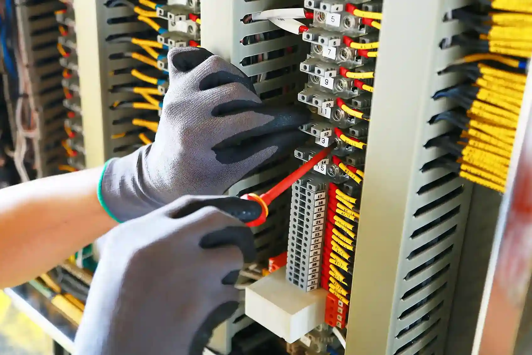 A close-up shot of a gloved hand using a screwdriver to connect a red wire to a terminal block inside an electrical panel.