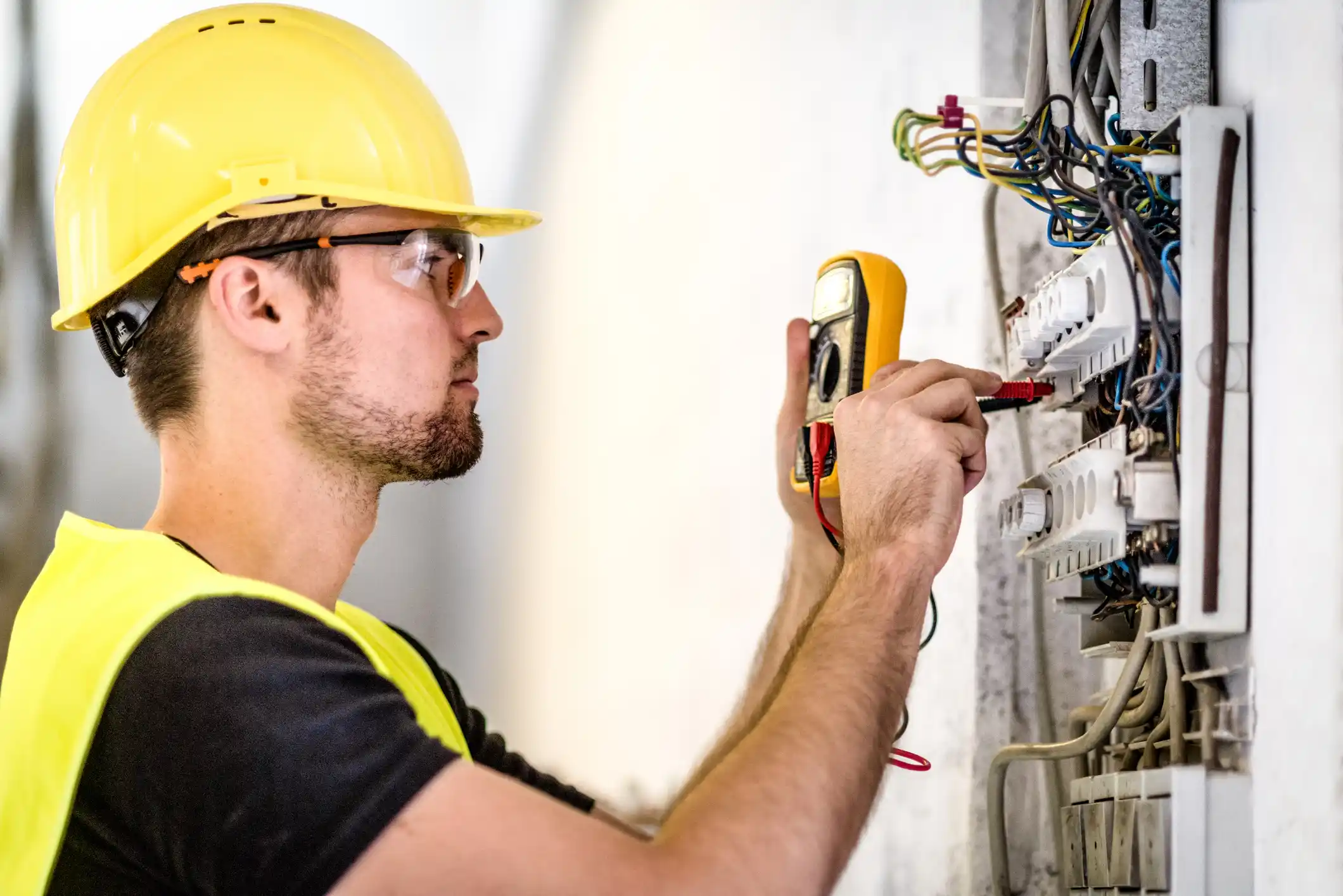 An electrician in a hard hat and safety glasses uses a multimeter to check the voltage on a fuse box.