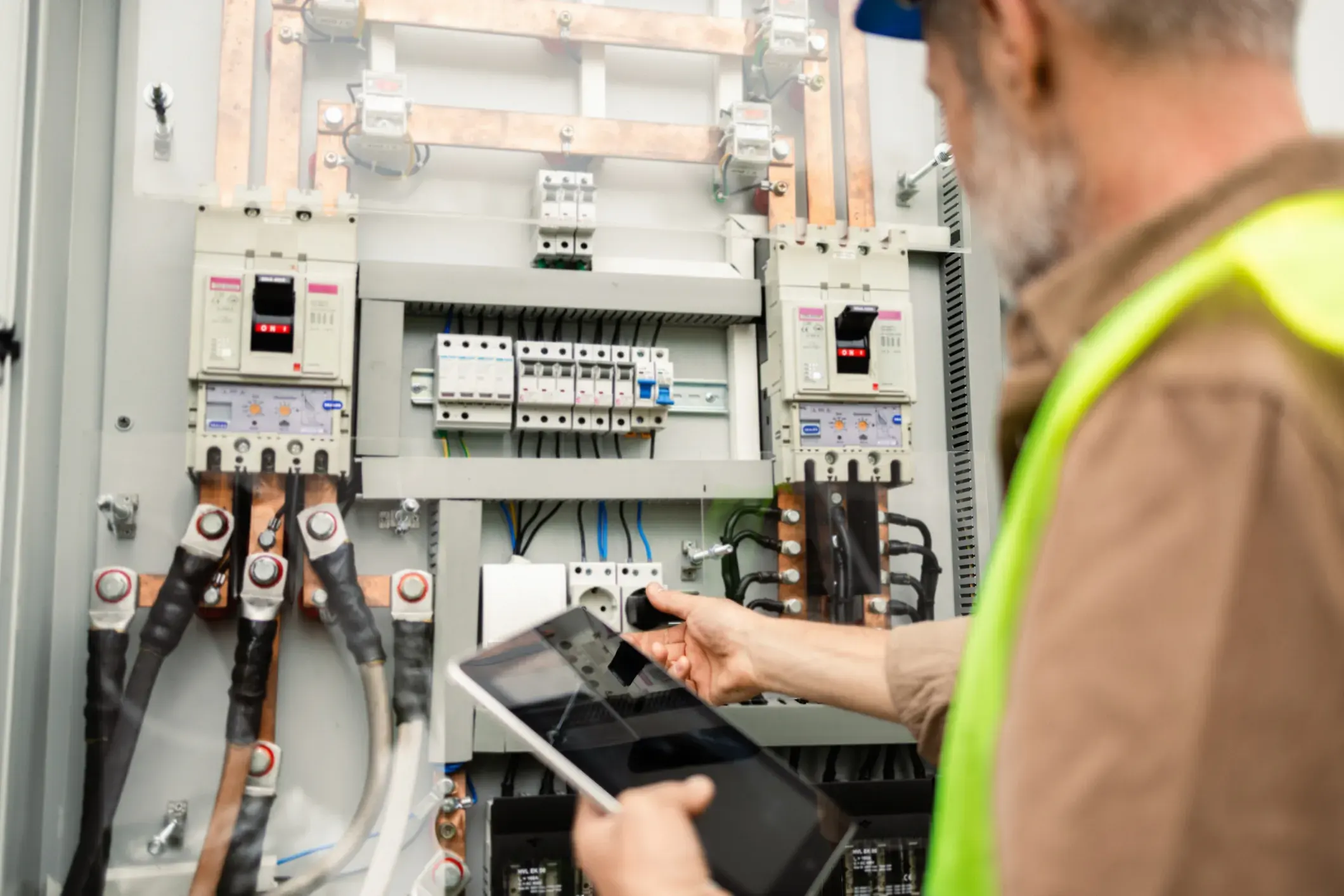 A technician with a beard and a hard hat uses a digital tablet while working on a large electrical panel with multiple circuit breakers and thick cables.