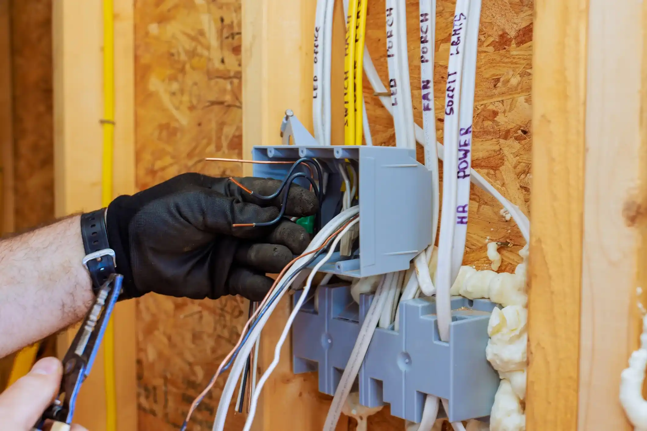 An electrician wearing a black glove uses wire strippers to work with a bundle of wires coming from a gray junction box on a wood-framed wall.