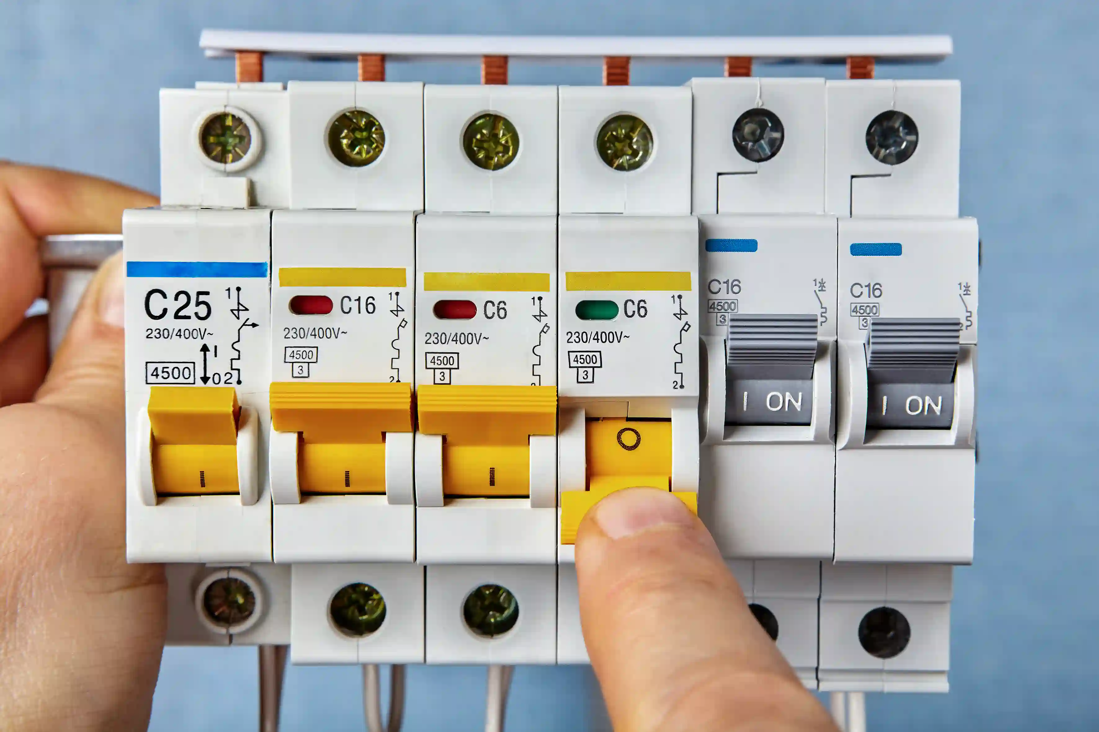 A close-up view of a hand pressing the yellow toggle switch of a modular circuit breaker in a row of several similar devices. The breakers are labeled with ratings like 'C25' and 'C16' and show copper busbars connecting them at the top.